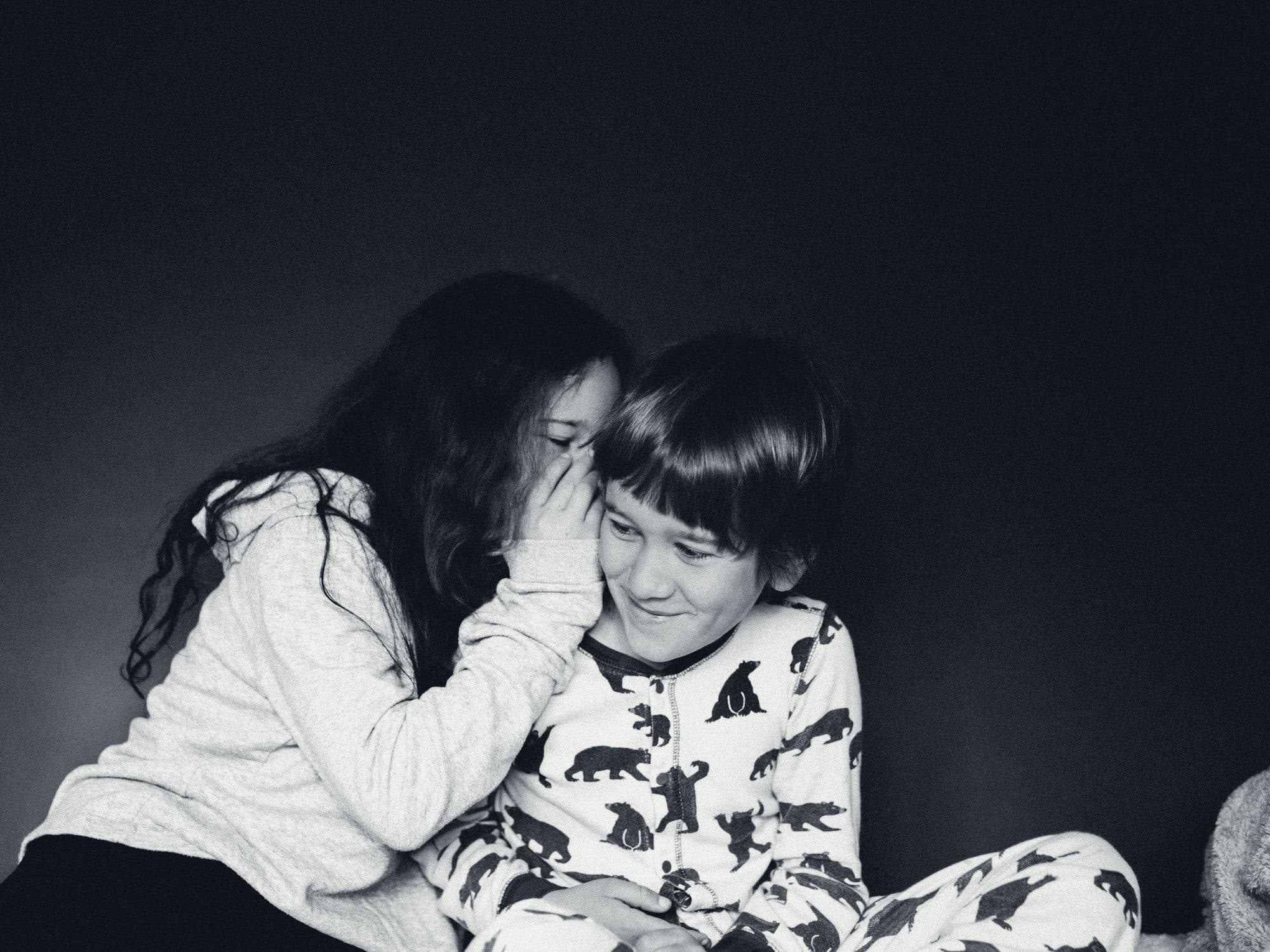 A girl whispers into a younger boy’s ear